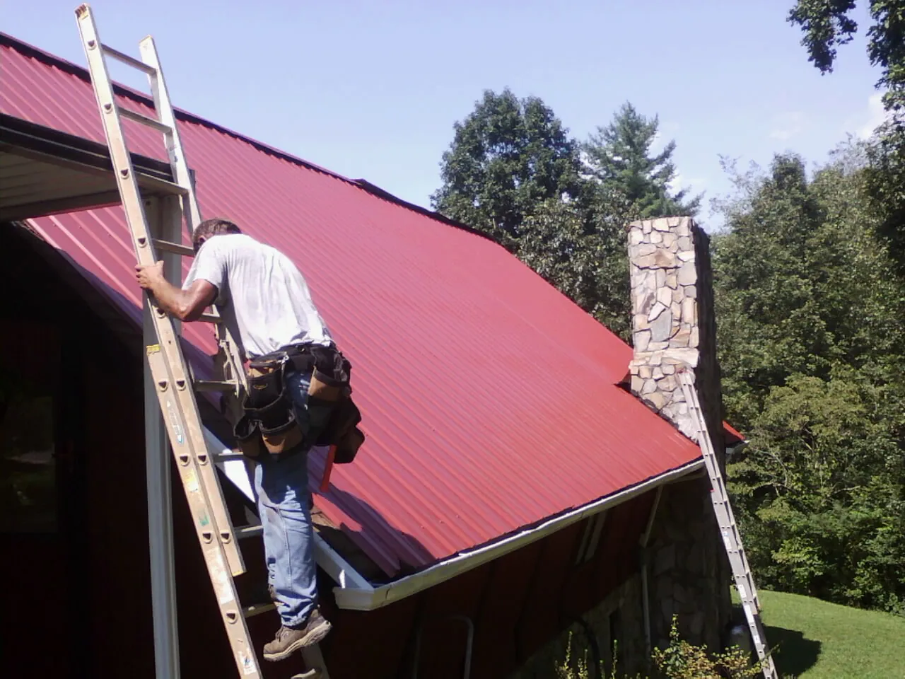Roofer on a ladder beside a red metal roof and stone chimney — Soffit & Fascia Repair by Roofing Brothers in Bronx